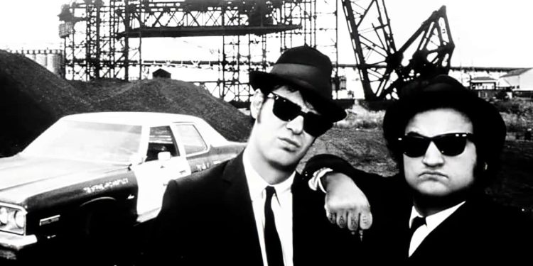 Jake and Elwood Blues from "The Blues Brothers" posing with the Bluesmobile in a black and white photo.